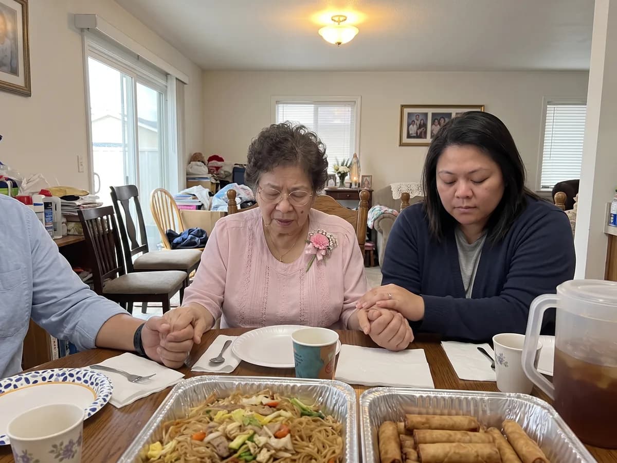A grandmother with her eyes closed holding hands with her daughter to say grace before her 70th birthday lunch, foil trays of pancit and lumpia on the table