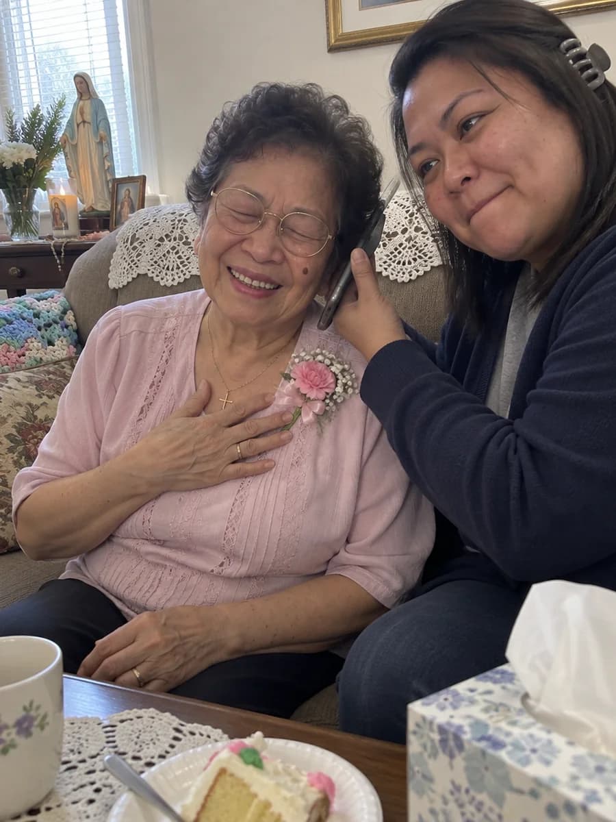 A grandmother in her church blouse and corsage with her hand over her heart, eyes closed and smiling, as her daughter holds a phone to her ear to play her a song
