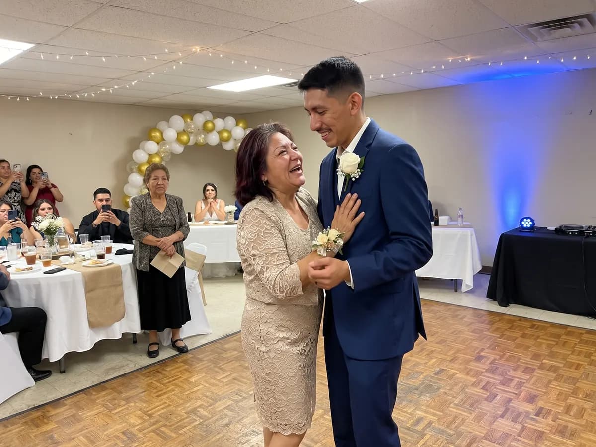 A groom slow dancing with his mother at the reception as she laughs and cries in surprise, guests filming and an aunt waiting with a napkin at the edge of the floor