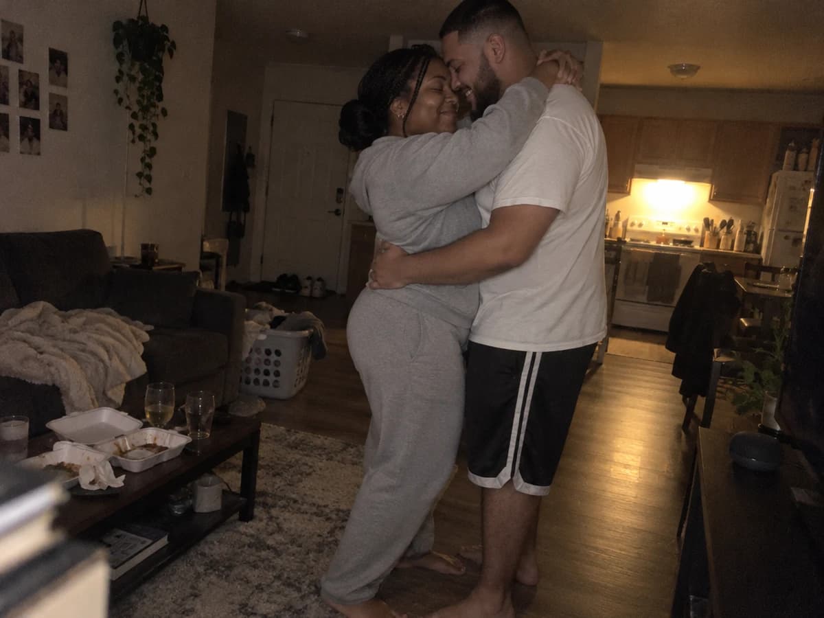 A couple slow dancing barefoot in their dim living room at night, foreheads together, takeout and two glasses on the coffee table