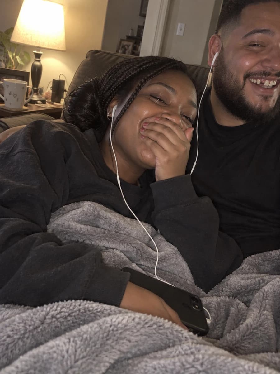 A woman under a blanket on the couch covering her mouth with happy tears as she listens to a song through one earbud, her partner sharing the other and laughing