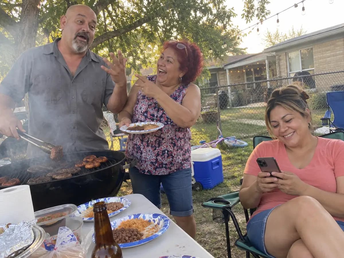A man at the grill telling a story at a backyard barbecue while his sister laughs and corrects him and his wife quietly types notes on her phone