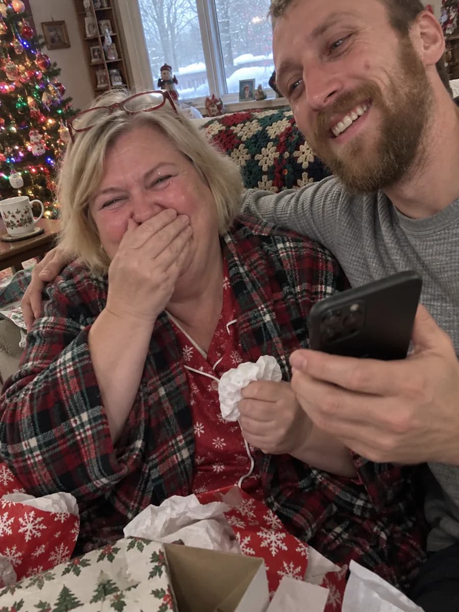 A mother in a flannel robe covering her mouth with happy tears on Christmas morning as her son, arm around her, plays her a song on his phone