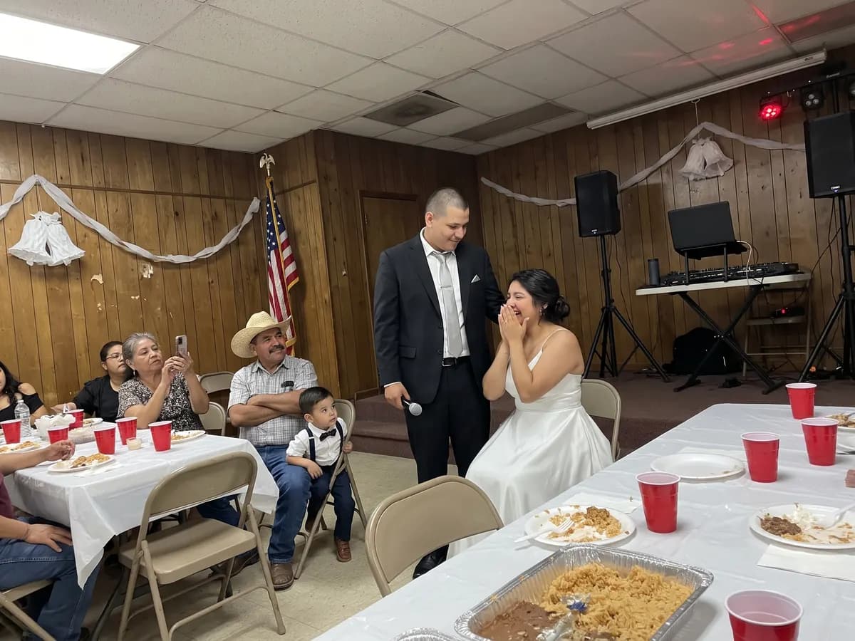 A groom in a black suit standing beside his bride at their VFW hall reception as she covers her mouth, laughing and teary, while guests watch and film