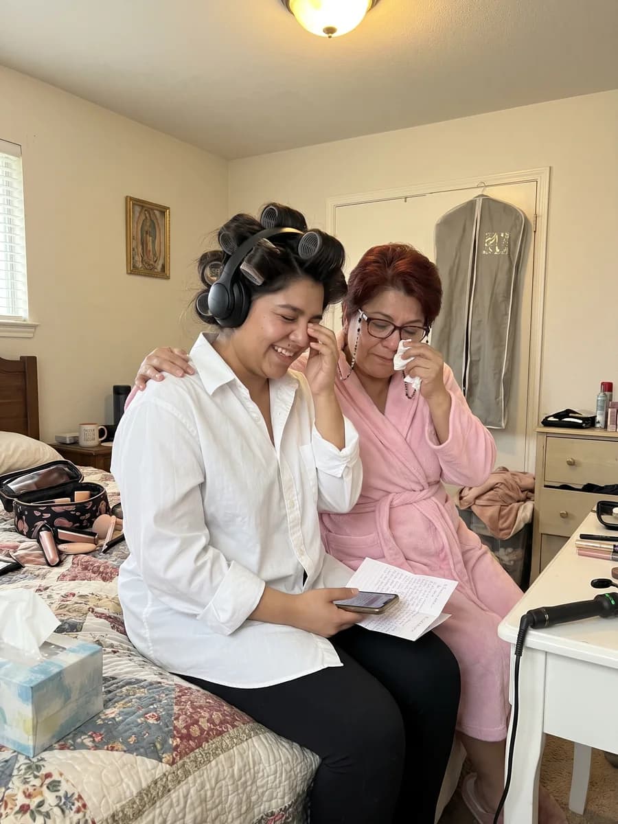 A bride in hair rollers and headphones holding a handwritten note, laughing through happy tears as her mother hugs her on the morning of the wedding