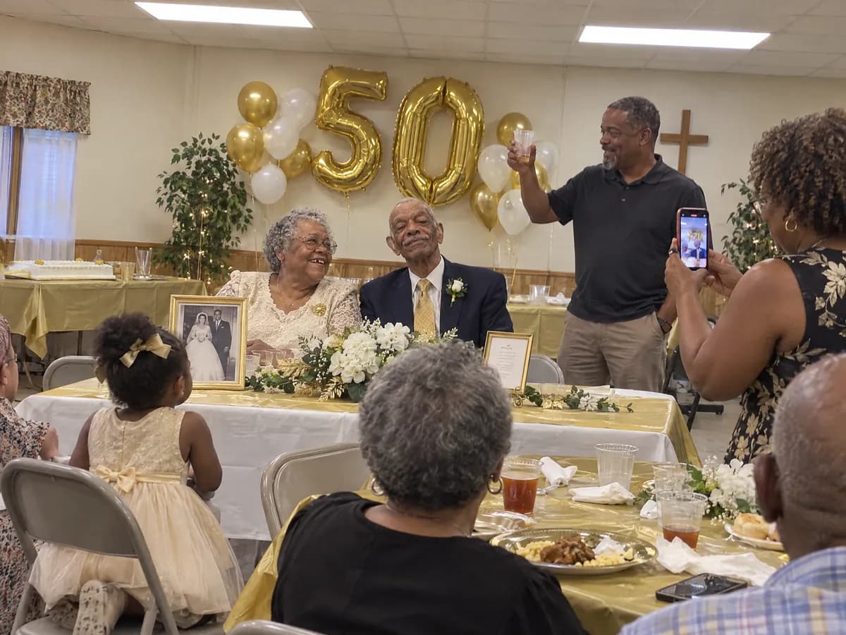An older couple at the head table of their 50th anniversary party, gold 50 balloons behind them and their wedding photo in front, as their son raises a toast and a relative films
