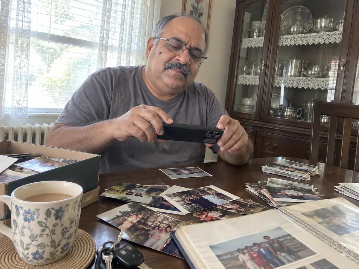 A man at his dining table photographing old printed snapshots with his phone in window daylight, a shoebox and an open photo album spread out around him