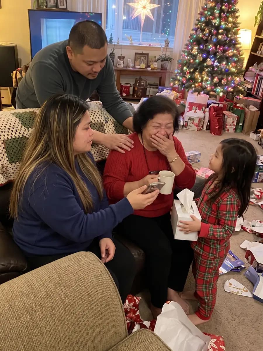 A grandmother on the couch on Christmas evening, hand over her mouth and crying happy tears, as her daughter plays a song on her phone and a granddaughter brings tissues