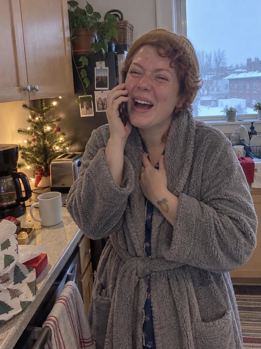 A woman in a robe in her apartment kitchen on Christmas morning, phone to her ear, laughing and crying as she listens to a song