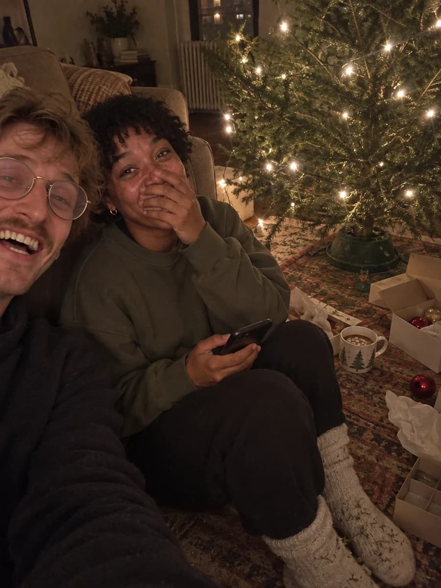 A young woman on the floor by a Christmas tree lit only by its string lights, covering her mouth with happy tears as her partner laughs beside her