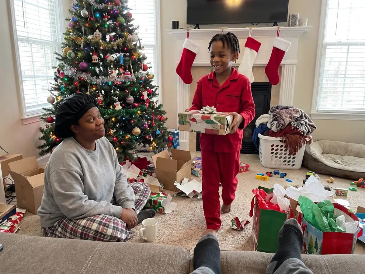 A boy in red pajamas carrying a wrapped shoebox across the living room to his mom by the Christmas tree on Christmas morning